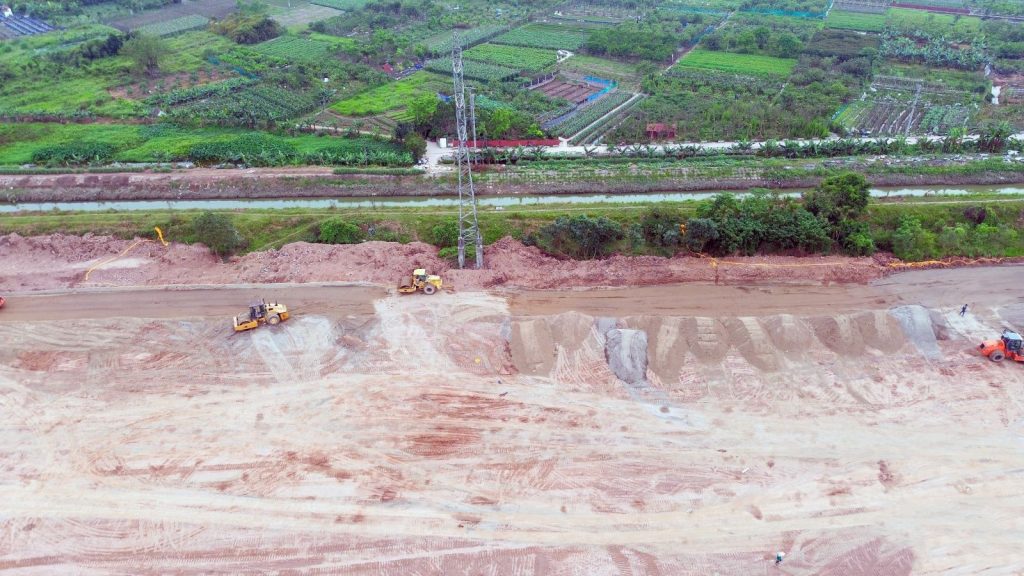 Máy ủi và lu làm việc trên công trường san lấp đất tại dự án Sunshine Grand City, phía sau là kênh nước và khu vực trồng trọt xanh mát.