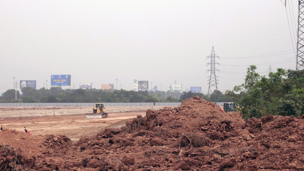 Ảnh cận cảnh công trường với máy ủi đang làm việc, các biển quảng cáo và cột điện cao thế phía xa, xung quanh là đống đất và cây xanh.